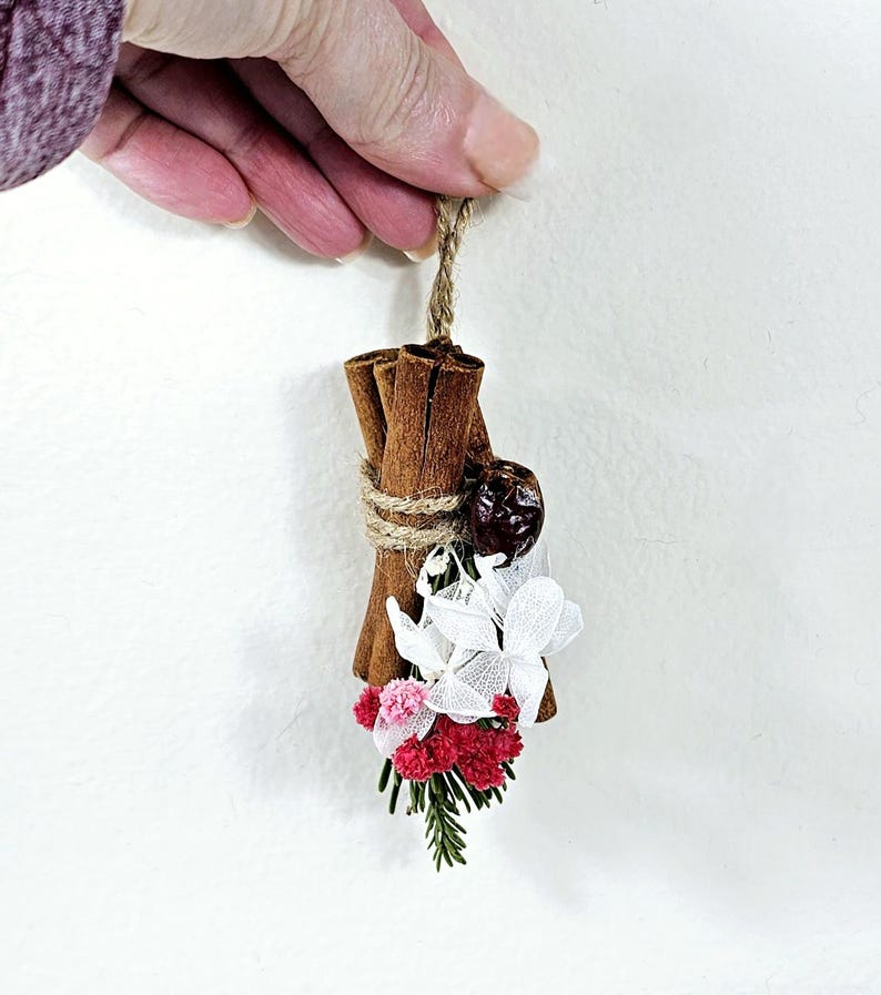 Puede incluir: Un manojo decorativo de palitos de canela atados con hilo, adornado con flores secas blancas, rosas y rojas, y una baya oscura. Cuelga de una cuerda de fibra natural, creando un objeto decorativo r&uacute;stico.