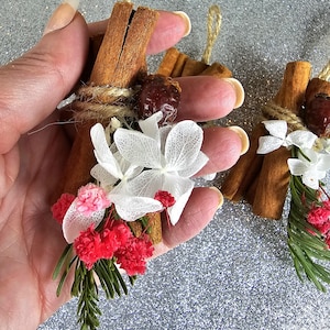 Puede incluir: Adornos de palitos de canela adornados con flores blancas secas, flores rojas y rosas y una semilla marr&oacute;n. Los palitos de canela est&aacute;n atados con hilo y tienen un lazo de hilo natural para colgar. El fondo es plateado brillante.