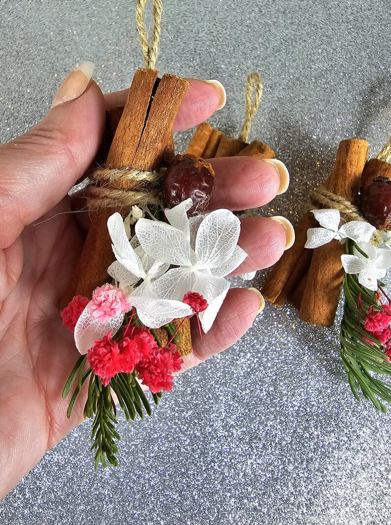 Puede incluir: Adornos de palitos de canela adornados con flores blancas secas, flores rojas y rosas y una semilla marr&oacute;n. Los palitos de canela est&aacute;n atados con hilo y tienen un lazo de hilo natural para colgar. El fondo es plateado brillante.