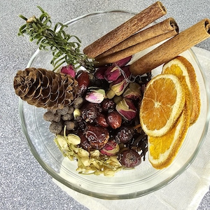 May include: A clear glass bowl filled with dried ingredients for potpourri. The bowl contains cinnamon sticks, dried orange slices, rosebuds, a pine cone, and other spices. The arrangement sits on a white cloth.