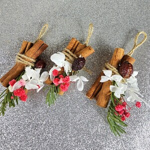 Puede incluir: Tres paquetes decorativos con palitos de canela, bayas secas, flores blancas y rosas y vegetaci&oacute;n. Cada paquete est&aacute; atado con hilo y tiene un lazo para colgar. El fondo es una superficie plateada brillante.
