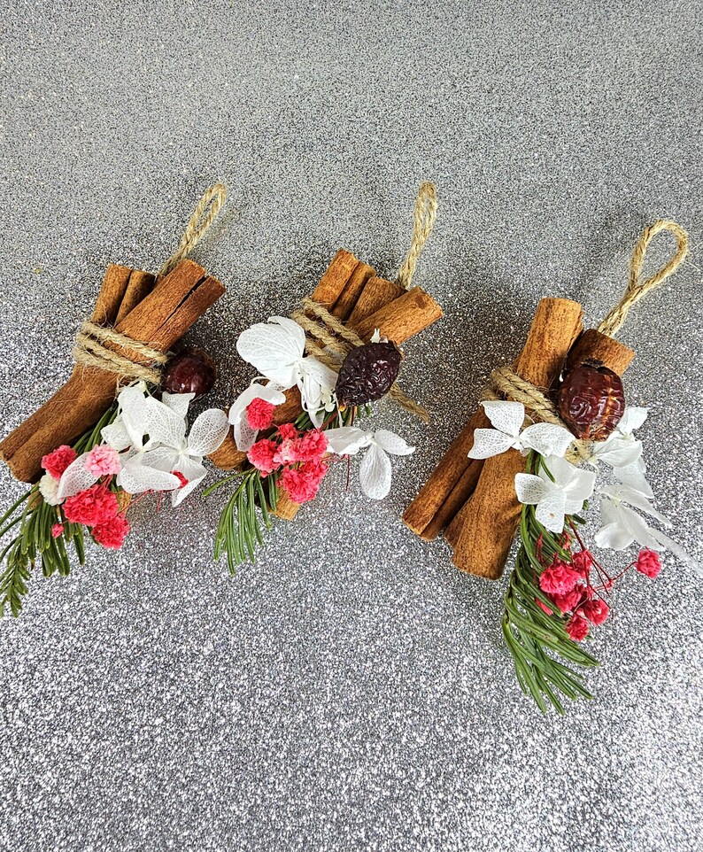 Puede incluir: Tres paquetes decorativos con palitos de canela, bayas secas, flores blancas y rosas y vegetaci&oacute;n. Cada paquete est&aacute; atado con hilo y tiene un lazo para colgar. El fondo es una superficie plateada brillante.