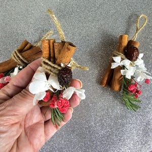Puede incluir: Primer plano de tres paquetes decorativos con palitos de canela, atados con hilo y adornados con flores blancas, bayas rojas y vegetaci&oacute;n. Cada paquete tiene un lazo de hilo para colgar. El fondo es una superficie plateada brillante.