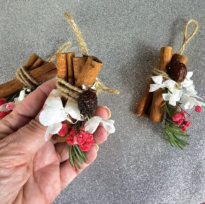 Puede incluir: Primer plano de tres paquetes decorativos con palitos de canela, atados con hilo y adornados con flores blancas, bayas rojas y vegetaci&oacute;n. Cada paquete tiene un lazo de hilo para colgar. El fondo es una superficie plateada brillante.