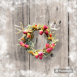 May include: A circular wreath with pink rose buds, green foliage, and gold-coloured accents. The wreath has small, dried berries and gold-coloured antlers. It is set against a rustic, grey wooden background.