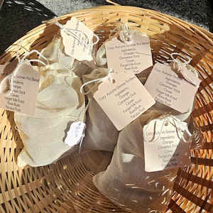 May include: A woven basket holds several beige mesh bags, each with a tag that reads "Cozy Autumn Simmer Pot Blend." The tags list ingredients like oranges, cinnamon sticks, and cloves. The basket is made of light brown wood.