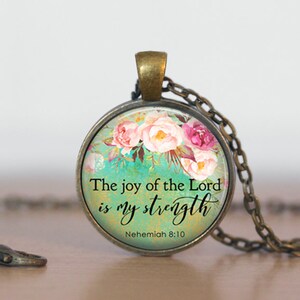 Puede incluir: Un collar con un colgante redondo de vidrio con un dise&ntilde;o floral y el texto "The joy of the Lord is my strength" y "Nehemiah 8:10".