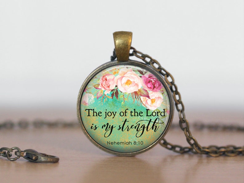 Puede incluir: Un collar con un colgante redondo de vidrio con un dise&ntilde;o floral y el texto "The joy of the Lord is my strength" y "Nehemiah 8:10".