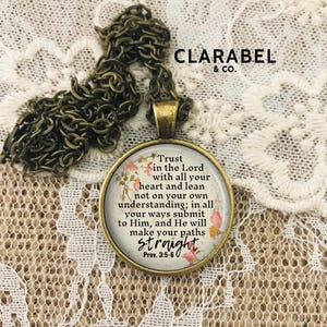 May include: A gold necklace with a round glass pendant featuring a floral design and the text "Trust in the Lord with all your heart and lean not on your own understanding; in all your ways submit to Him, and He will make your paths straight Prov. 3:5-6".