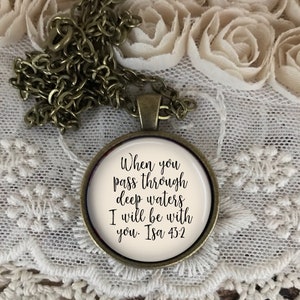 May include: A bronze necklace with a round pendant featuring the quote "When you pass through deep waters I will be with you. Isa 43:2" in black cursive lettering on a white background.