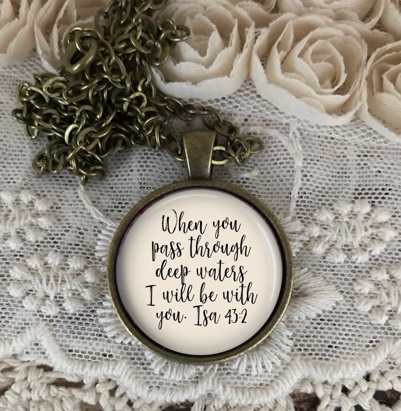 May include: A bronze necklace with a round pendant featuring the quote "When you pass through deep waters I will be with you. Isa 43:2" in black cursive lettering on a white background.