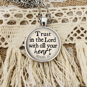 May include: A silver-toned necklace with a round pendant. The pendant features the text "Trust in the Lord with all your heart" in black, with "Proverbs 3:5" below. The necklace rests on a cream-colored, textured surface.
