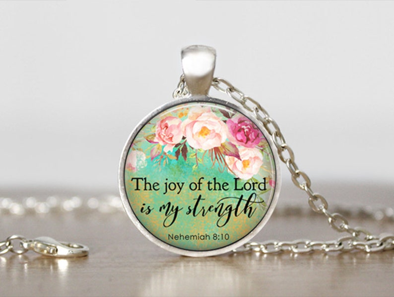 Puede incluir: Un collar de plata con un colgante redondo de vidrio con un dise&ntilde;o floral y la cita "La alegr&iacute;a del Se&ntilde;or es mi fortaleza" con la referencia b&iacute;blica "Nehem&iacute;as 8:10".