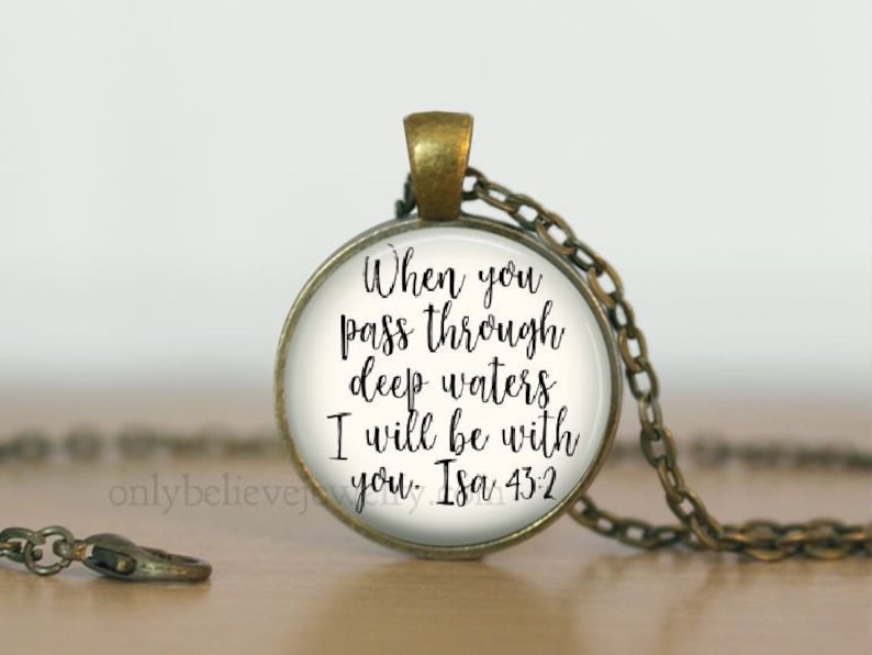 May include: A silver necklace with a round glass pendant featuring the text "When you pass through deep waters I will be with you. Isa 43:2" in black cursive lettering.