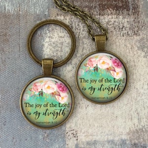 Puede incluir: Un llavero y un collar de bronce con un colgante redondo de vidrio que presenta un dise&ntilde;o floral y la cita "La alegr&iacute;a del Se&ntilde;or es mi fortaleza" con la referencia b&iacute;blica "Nehem&iacute;as 8:10".