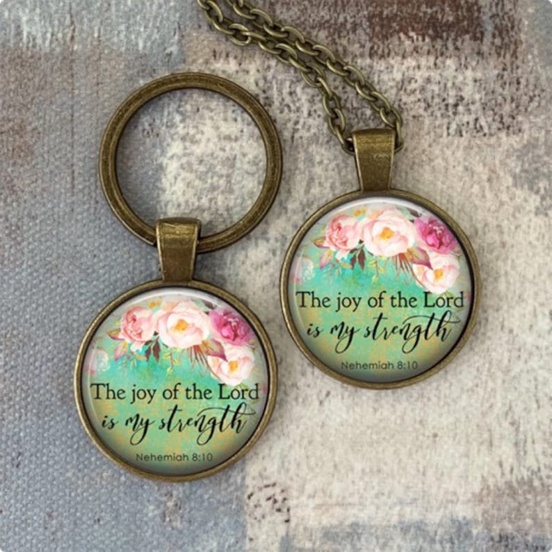 Puede incluir: Un llavero y un collar de bronce con un colgante redondo de vidrio que presenta un dise&ntilde;o floral y la cita "La alegr&iacute;a del Se&ntilde;or es mi fortaleza" con la referencia b&iacute;blica "Nehem&iacute;as 8:10".