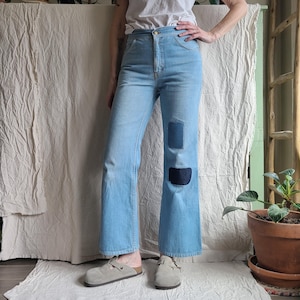 May include: Light blue high-waisted denim jeans featuring a straight-leg cut and two dark blue rectangular patches on the left leg. The jeans have a classic button and zipper fly closure, visible stitching, and a clean hemline at the ankles.