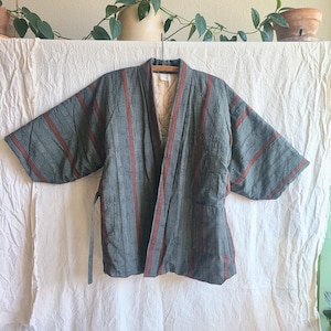 May include: A quilted jacket featuring a dark green and rust-colored striped pattern with subtle embroidered floral details. The garment has wide sleeves, an open front, and a beige quilted interior lining, hanging on a wooden hanger.