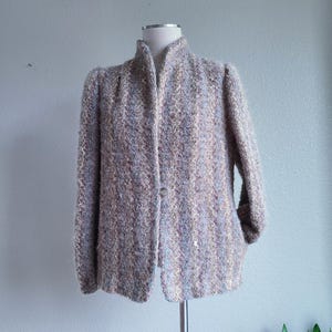 May include: A multi-colored, textured jacket with a high collar and long sleeves. The jacket is displayed on a mannequin and features a single button closure. The fabric has a mix of colors, including beige, blue, and gold.