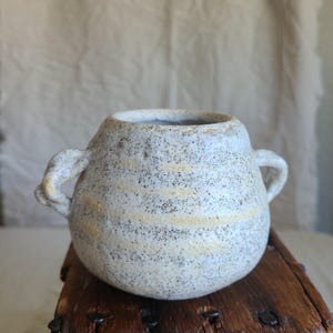 May include: A small, speckled ceramic vase with two curved handles. The vase has a rounded body and a wide opening. The exterior features a cream-colored base with black speckles and horizontal bands of a lighter cream color.