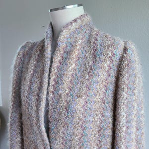 May include: A textured, multi-colored jacket with a high collar. The jacket is displayed on a mannequin and features a blend of cream, lavender, and teal hues. The fabric has a fuzzy, bouclé-like texture, suggesting a warm, cozy garment.