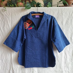 May include: A dark blue denim pullover shirt featuring a V-neckline, wide three-quarter length sleeves, and a curved hem. The upper chest area displays an embroidered patch with a red chili pepper, a striped circular motif, and geometric shapes.