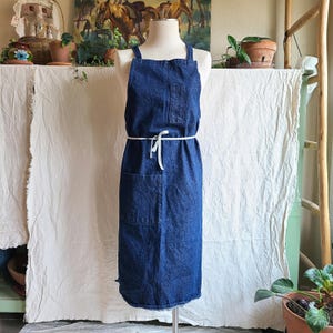May include: A dark blue denim apron featuring a rectangular chest pocket and a large lower pocket. The apron is secured at the waist with a thin white tie and has two shoulder straps that cross at the back, displayed on a white mannequin torso.