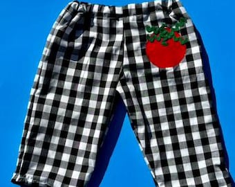 May include: Pants featuring a black and white gingham check pattern with an elastic waistband. A bright red tomato applique with green leafy stems is stitched onto the upper right side of the garment, adding a distinct visual detail.