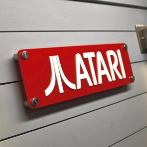 Puede incluir: Un letrero rojo con letras blancas que dice "ATARI" con un logotipo "A" estilizado. El letrero está montado en una pared de madera blanca.