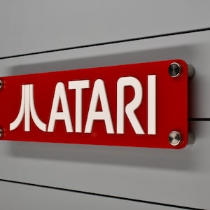 Puede incluir: Un letrero rectangular rojo con letras blancas que dice "ATARI" con un logotipo de Pac-Man estilizado a la izquierda del texto. El letrero está montado en una pared gris con tornillos plateados.