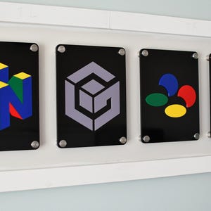 Könnte beinhalten: Eine Wandanzeige mit fünf schwarzen quadratischen Plaketten mit verschiedenen farbigen Videospiel-Logos. Die Logos sind für Nintendo 64, Nintendo GameCube, Super Nintendo und Nintendo Switch.