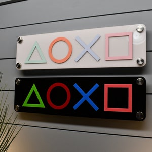 May include: Two rectangular signs with the PlayStation controller symbols. The top sign is white with light blue, light green, and orange symbols. The bottom sign is black with red, green, and blue symbols.