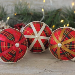 May include: Three Christmas ornaments made of red fabric with a plaid pattern and white or gold ribbon accents. Each ornament has a white pearl embellishment in the center.