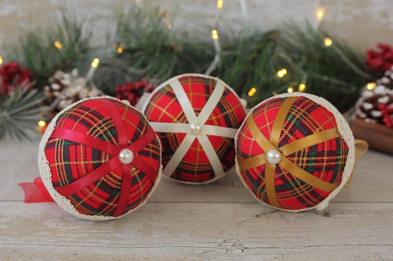 May include: Three Christmas ornaments made of red fabric with a plaid pattern and white or gold ribbon accents. Each ornament has a white pearl embellishment in the center.
