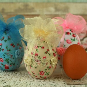 Peut inclure: Des &oelig;ufs de P&acirc;ques d&eacute;coratifs en tissu floral, dentelle et n&oelig;uds en ruban bleu, jaune et rose. Un seul &oelig;uf brun est &eacute;galement pr&eacute;sent. Les &oelig;ufs sont pos&eacute;s sur une surface en dentelle blanche, &eacute;voquant une ambiance festive.