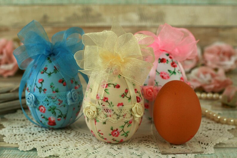 Peut inclure: Des &oelig;ufs de P&acirc;ques d&eacute;coratifs en tissu floral, dentelle et n&oelig;uds en ruban bleu, jaune et rose. Un seul &oelig;uf brun est &eacute;galement pr&eacute;sent. Les &oelig;ufs sont pos&eacute;s sur une surface en dentelle blanche, &eacute;voquant une ambiance festive.