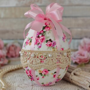 May include: An egg-shaped ornament with a floral pattern in pink and white. It features a beige lace band, faux pearls, and a pink satin bow. Pink satin ribbons are also attached. The ornament is a decorative item.