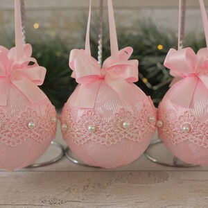 May include: Three pink Christmas baubles with white lace and pearl accents. Each bauble has a pink satin ribbon bow.