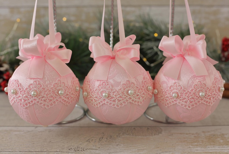 May include: Three pink Christmas baubles with white lace and pearl accents. Each bauble has a pink satin ribbon bow.