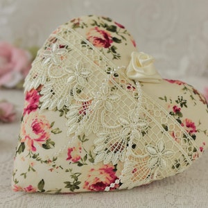 May include: A heart-shaped pillow with a floral print and lace trim. The pillow is cream-colored with pink and red roses.
