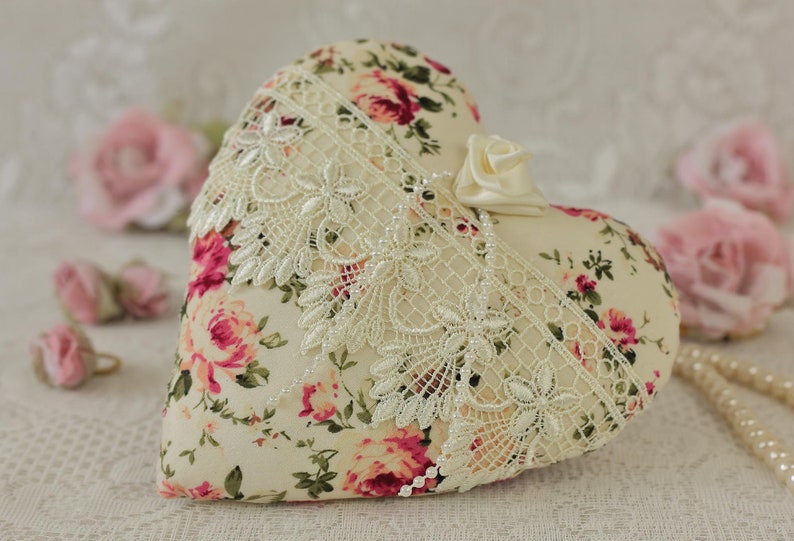 May include: A heart-shaped pillow with a floral print and lace trim. The pillow is cream-colored with pink and red roses.