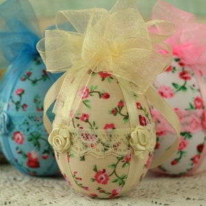 Peut inclure: Trois &oelig;ufs de P&acirc;ques d&eacute;coratifs en tissu. Les &oelig;ufs sont recouverts de motifs floraux avec des fleurs roses et rouges. Chaque &oelig;uf a un n&oelig;ud en ruban en haut et est orn&eacute; de dentelle et de petites roses. Les &oelig;ufs sont dans des tons de bleu, cr&egrave;me et rose.