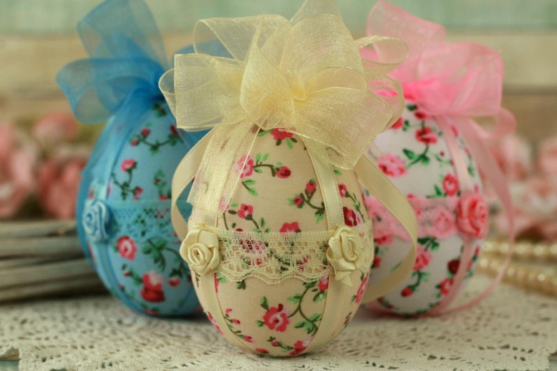 Peut inclure: Trois &oelig;ufs de P&acirc;ques d&eacute;coratifs en tissu. Les &oelig;ufs sont recouverts de motifs floraux avec des fleurs roses et rouges. Chaque &oelig;uf a un n&oelig;ud en ruban en haut et est orn&eacute; de dentelle et de petites roses. Les &oelig;ufs sont dans des tons de bleu, cr&egrave;me et rose.