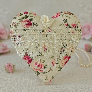 May include: A heart-shaped pillow with a floral print and lace trim. The pillow is decorated with a small white satin rose and is displayed on a white surface.
