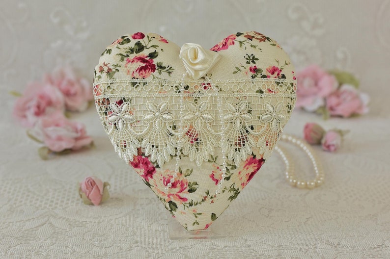 May include: A heart-shaped pillow with a floral print and lace trim. The pillow is decorated with a small white satin rose and is displayed on a white surface.
