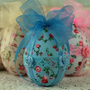 Peut inclure: Des &oelig;ufs de P&acirc;ques d&eacute;coratifs en tissu aux couleurs pastel. Les &oelig;ufs sont recouverts de motifs floraux et orn&eacute;s de rubans et de rosettes. L'&oelig;uf central est bleu clair avec des fleurs rouges, une garniture en dentelle et un n&oelig;ud bleu.