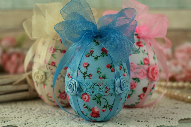 Peut inclure: Des &oelig;ufs de P&acirc;ques d&eacute;coratifs en tissu aux couleurs pastel. Les &oelig;ufs sont recouverts de motifs floraux et orn&eacute;s de rubans et de rosettes. L'&oelig;uf central est bleu clair avec des fleurs rouges, une garniture en dentelle et un n&oelig;ud bleu.