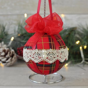 May include: Red and green plaid Christmas ornament with white lace trim and a red bow. The ornament is on a silver stand.