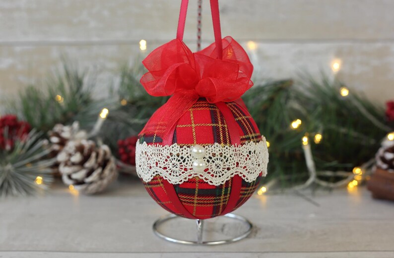May include: Red and green plaid Christmas ornament with white lace trim and a red bow. The ornament is on a silver stand.
