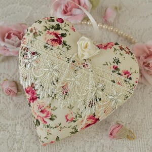 May include: A heart-shaped pillow with a floral print and lace trim. The pillow is on a white lace tablecloth with pink roses.